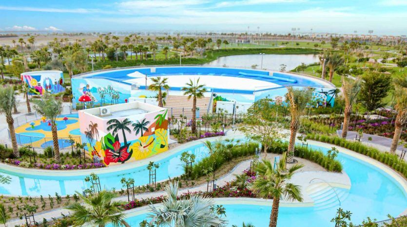 Vue aérienne d'un parc aquatique extérieur animé comprenant de grandes piscines ornées de peintures murales artistiques, entourées d'une verdure luxuriante et d'un lac pittoresque digne d'un investissement en arrière-plan.