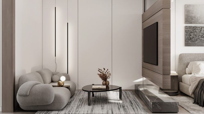 Un salon moderne dans un appartement de Dubaï comprenant un canapé gris moelleux et incurvé, une table basse ronde, un meuble de télévision élégant et un lit confortable en arrière-plan, le tout dans un espace aux tons neutres.