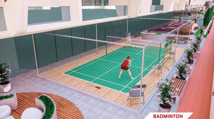 Une personne en tenue rouge jouant au badminton sur un terrain vert dans un appartement sur le toit de Dubaï, entouré de barrières de verre, de plantes et de mobilier d'extérieur, avec des bâtiments en arrière-plan.