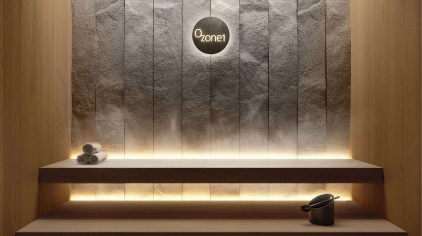 Un mur moderne et minimaliste dans une villa de Dubaï avec une surface texturée, éclairé par des lumières chaudes et apaisantes, comprenant une simple étagère en bois avec deux serviettes roulées et une tasse sombre. Un panneau rond et éclairé indiquant « Zone1 » est fixé au mur.