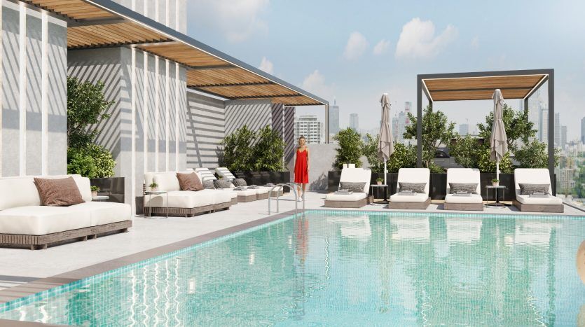 Un espace piscine sur le toit avec des chaises longues et des cabanes modernes, décorées de plantes. Une personne vêtue d’une robe rouge se tient au bord de la piscine et contemple le paysage urbain de Dubaï.