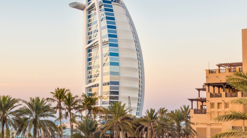 Vue de l'hôtel Burj Al Arab à Dubaï au coucher du soleil, encadrée par des palmiers et l'architecture de la villa adjacente de Dubaï, sous un ciel rose tendre.