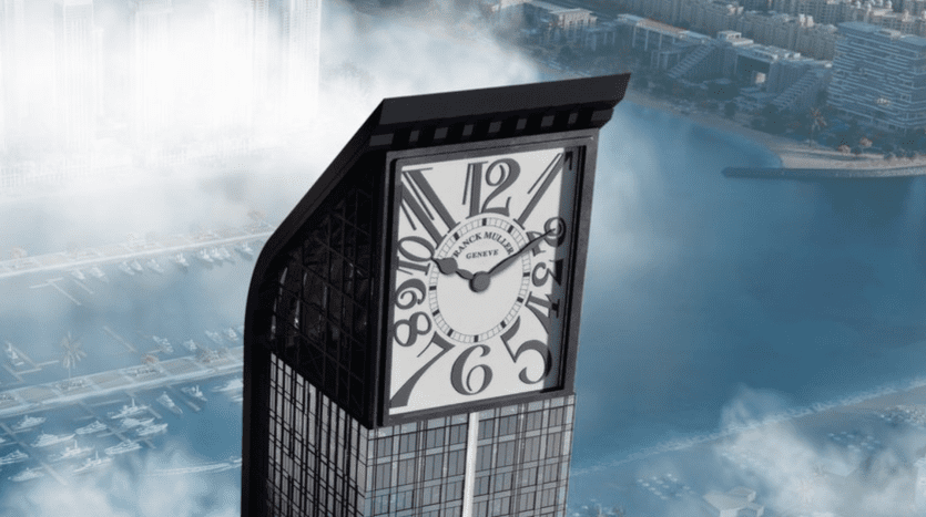 Un immeuble de grande hauteur avec un grand cadran d'horloge intégré à son sommet, surplombant une ville côtière de Dubaï partiellement obscurcie par le brouillard matinal.