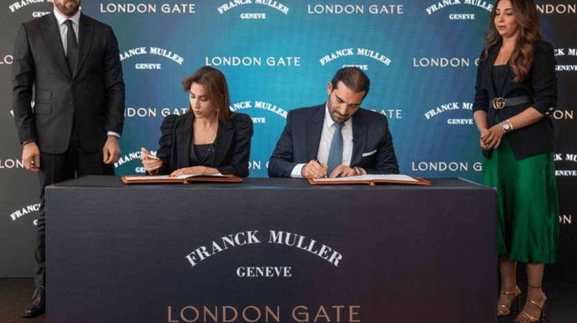 Deux personnes signent des documents à une table avec « Agence Immobilière Dubaï » sur le devant. Ils sont flanqués de deux individus debout dans un contexte professionnel.