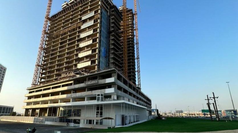 Un grand bâtiment en construction à Dubaï avec plusieurs étages et des échafaudages, sur fond de ciel dégagé. Le premier plan montre une zone herbeuse et d'autres bâtiments sont visibles au loin.