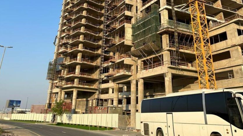 Un bâtiment à plusieurs étages en construction, avec des échafaudages visibles et une grue à côté. Un bus blanc est garé sur une route au premier plan, sous un ciel bleu clair. Ce site représente l'un des derniers projets gérés par une agence immobilière leader à Dubaï.