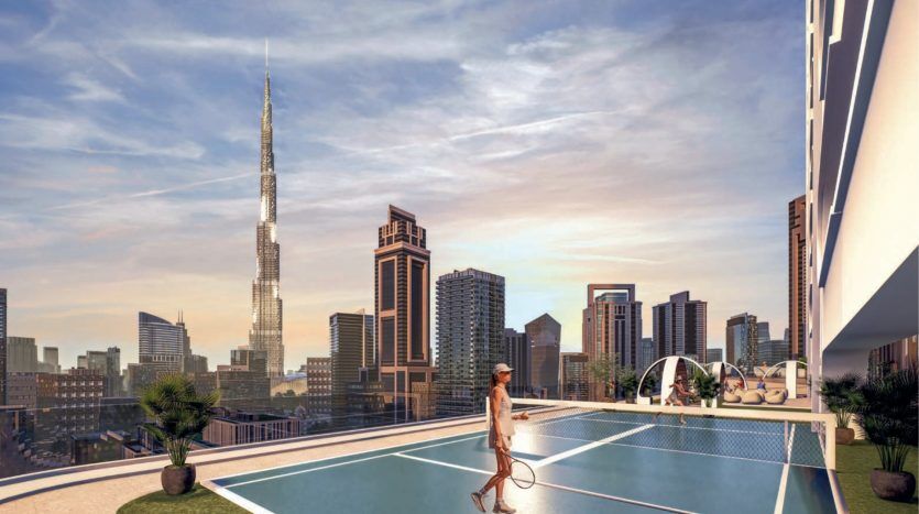 A person plays tennis on a rooftop court in a modern cityscape at sunset in Dubai, with tall buildings and a particularly striking skyscraper in the background.