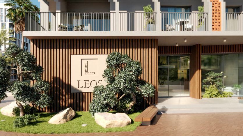 Extérieur d'un immeuble d'appartements moderne à Dubaï avec une verdure luxuriante, présentant un design en bois et en métal avec une pancarte indiquant "LEOS", situé sous un balcon dans un cadre ensoleillé.