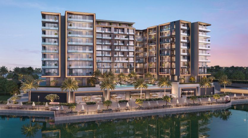 Un immeuble résidentiel moderne en bord de mer à Dubaï au crépuscule, doté de balcons, de grandes fenêtres et d'un aménagement paysager luxuriant, se reflétant doucement dans l'eau calme en contrebas.