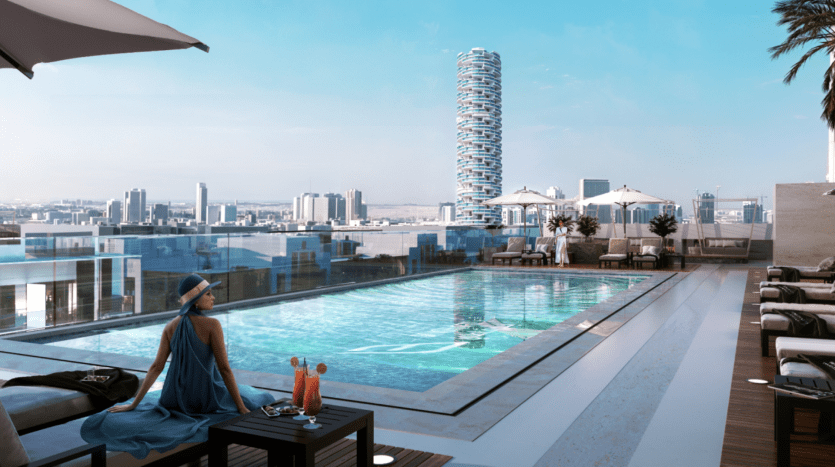 Une femme vêtue d'une robe bleue est assise au bord d'une piscine à débordement sur un toit, surplombant un paysage urbain avec des gratte-ciel modernes à Dubaï. Une boisson rafraîchissante est posée sur une table à côté d'elle.