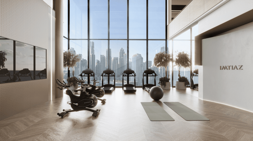 Une salle de sport moderne avec des tapis roulants et des vélos d'exercice faisant face à de grandes fenêtres présentant les toits de la ville de Dubaï. Il y a deux tapis de yoga et un ballon d'exercice sur un parquet.
