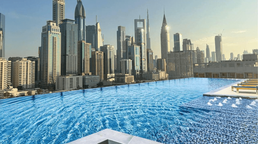 Piscine à débordement située dans un immeuble de grande hauteur à Dubaï avec une vue panoramique sur les toits d'une ville moderne et animée avec plusieurs gratte-ciel sous un ciel bleu clair.