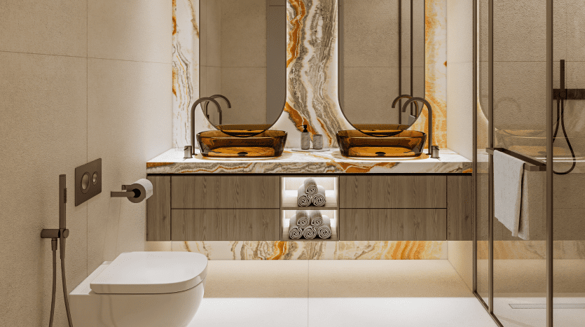 Une salle de bains moderne dans une villa de Dubaï comprenant deux vasques en verre ambré posées sur un comptoir en marbre, un grand miroir, des murs carrelés beiges et une pile de serviettes blanches soigneusement pliées.