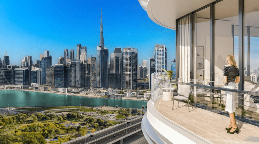 Une femme se tient sur un balcon spacieux de son appartement de Dubaï, surplombant un paysage urbain animé composé de gratte-ciel modernes et d’une grande étendue d’eau sous un ciel bleu clair.