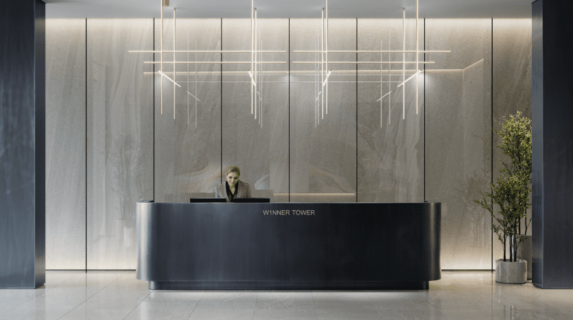 Un hall d'entrée d'hôtel moderne comprenant un comptoir de réception élégant avec une réceptionniste, des murs en marbre rétroéclairés et d'élégants luminaires linéaires, situé dans la Winner Tower. Une plante en pot ajoute une touche de verdure à cet espace qui rappelle une luxueuse villa de Dubaï.