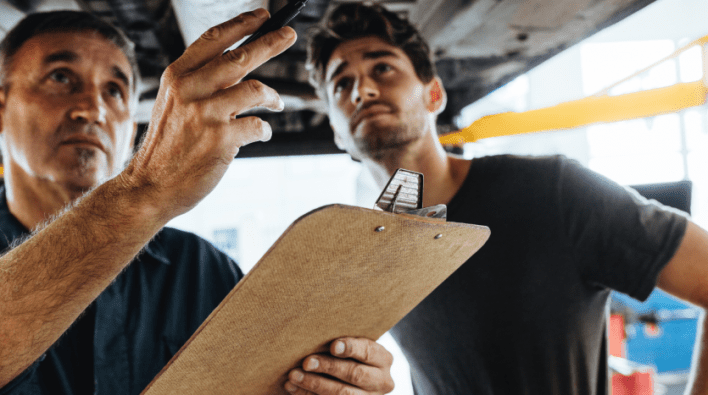 Deux hommes dans un atelier, un plus âgé et un plus jeune, inspectent une pièce automobile. L'homme plus âgé tient un presse-papier tandis que le plus jeune pointe vers le haut, tous deux attentivement concentrés.