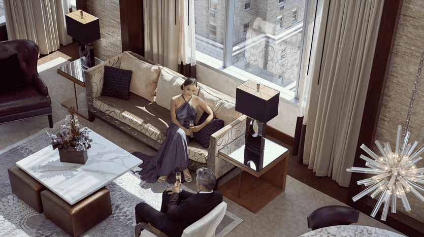 Un couple interagit dans un salon luxueux doté de grandes fenêtres, d'une décoration élégante et d'une vue sur la ville de Dubaï. La femme, assise et vêtue d’une robe fluide, s’adresse à l’homme en costume agenouillé à côté d’elle.
