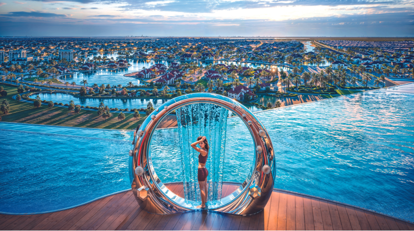Un homme se tient dans une structure métallique circulaire surmontée d’eau en cascade, donnant sur un complexe luxueux avec des bâtiments ornés et de vastes piscines bleues sous un ciel de coucher de soleil vibrant à Dubaï.