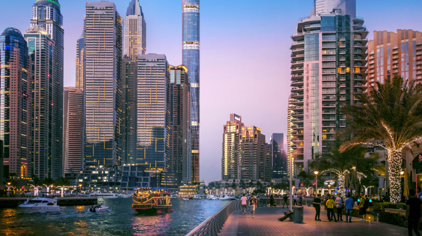 Une scène de front de mer animée au crépuscule avec des gratte-ciel modernes, une promenade animée et des bateaux sur l'eau, avec des gens marchant et profitant de la vue sur l'immobilier Dubaï.