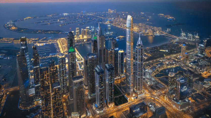 Vue aérienne de Dubaï au crépuscule, mettant en vedette des gratte-ciel illuminés, des routes très fréquentées et un paysage côtier avec des îles artificielles.