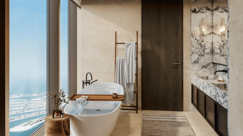 Une salle de bains luxueuse avec une baignoire autoportante, des accents de marbre et une grande fenêtre offrant une vue sur la mer dans un appartement exquis à Dubaï. Il y a une échelle en bois pour suspendre les serviettes et un tapis marron au sol.