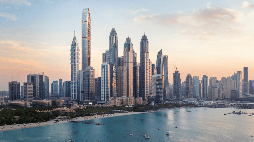 Une vue panoramique sur les toits d'une ville moderne au coucher du soleil, avec de grands gratte-ciel aux fenêtres lumineuses à côté d'un plan d'eau calme avec des bateaux et un premier plan d'arbres verts luxuriants à Dubaï.