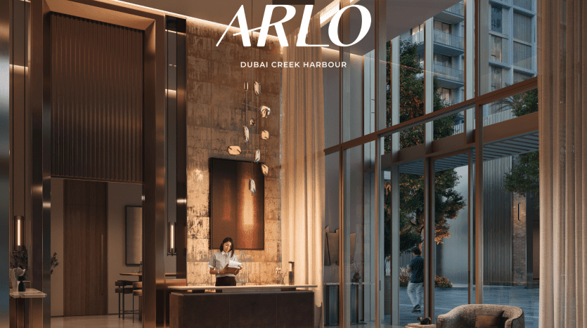 Un hall d'hôtel luxueux à Arlo Dubai Creek Harbour par Emaar. Un membre du personnel se tient derrière un comptoir de réception moderne et élégant, concentré sur son travail. L'espace comprend de hautes fenêtres en verre, un éclairage chaleureux, une décoration contemporaine et un coin salon extérieur tranquille visible à travers le verre.
