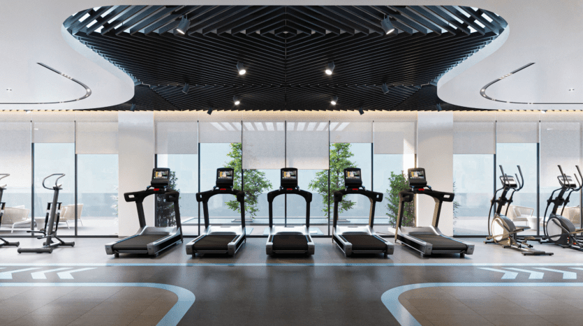 Une salle de sport moderne des résidences Azura comprend une rangée de cinq tapis roulants au centre, flanqués de divers appareils de fitness. De grandes baies vitrées en arrière-plan apportent de la lumière naturelle et offrent une vue sur la verdure extérieure. Le plafond présente un design géométrique noir élégant.