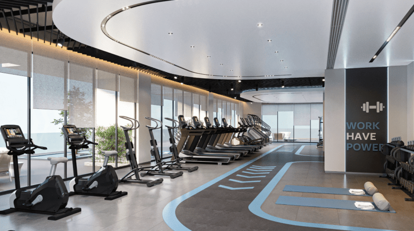 Une salle de sport moderne aux résidences Azura avec de grandes fenêtres, dotée d'une rangée de tapis roulants, de vélos d'exercice et d'appareils elliptiques. Les murs portent un texte de motivation : « WORK HAVE POWER ». La pièce est bien éclairée avec des éléments de design élégants et contemporains et un agencement propre et spacieux.