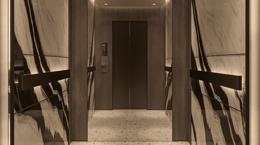 L'intérieur d'un ascenseur élégant et moderne du Primero Residences Al Furjan présente des panneaux en bois sombre et des surfaces en miroir. Le sol est recouvert de carreaux de marbre blanc aux subtiles veines grises. Un éclairage doux et ambiant crée une atmosphère luxueuse.