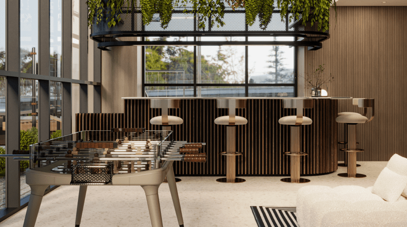 Coin salon moderne du Primero Residences Al Furjan comprenant un bar élégant avec des boiseries et des tabourets hauts, une table de baby-foot élégante avec un plateau en verre au premier plan et un canapé en forme de L avec un tapis à rayures. De grandes fenêtres offrent une vue sur la verdure, renforçant l'ambiance lumineuse et aérée de l'espace.