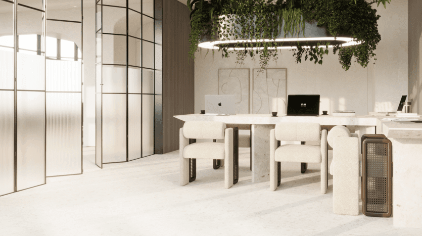 Un espace de bureau moderne au sein de Primero Residences Al Furjan comprend des chaises beiges, une grande table beige, deux ordinateurs portables et des plantes suspendues au-dessus dans un élégant luminaire noir. Des cloisons en verre divisent l'espace, avec des œuvres d'art minimalistes encadrées au mur. La lumière naturelle pénètre à travers de grandes fenêtres givrées.