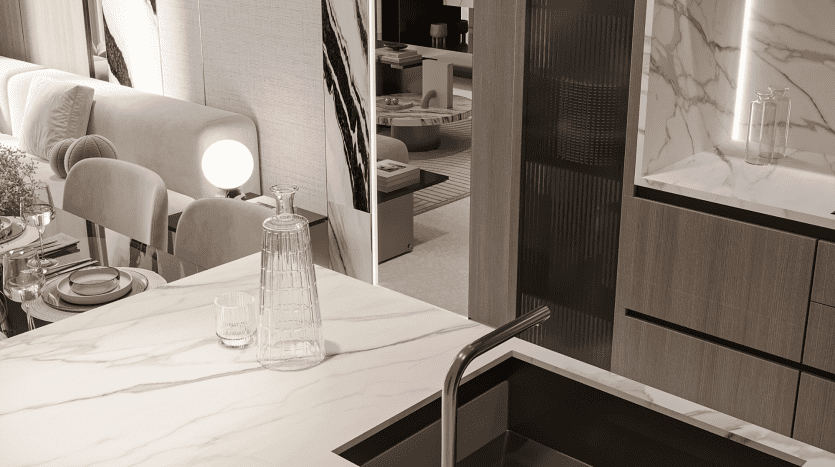 La cuisine et la salle à manger modernes du Primero Residences Al Furjan comprennent un comptoir en marbre avec une carafe à eau en verre et un verre, un robinet élégant, des chaises rembourrées beiges et un dosseret en marbre. L'espace suit une palette de couleurs neutres et luxueuses avec des accents de bois et un éclairage subtil.