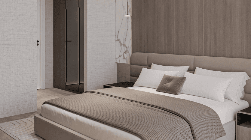 Une chambre moderne aux tons neutres du Primero Residences Al Furjan comprend un grand lit avec une literie blanche et beige et un oreiller décoratif. La tête de lit rembourrée est assortie au lit, tandis qu'une élégante armoire avec portes miroir est visible dans le coin, incarnant un design contemporain et minimaliste.