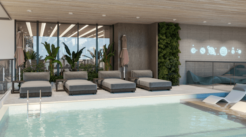 La piscine intérieure du Primero Residences Al Furjan comprend des chaises longues modernes alignées le long du bord, certaines avec des parasols. Des plantes vertes et un mur de jardin vertical ajoutent une touche de nature. Les grandes fenêtres apportent de la lumière naturelle, tandis que les néons contribuent à l'ambiance sereine.