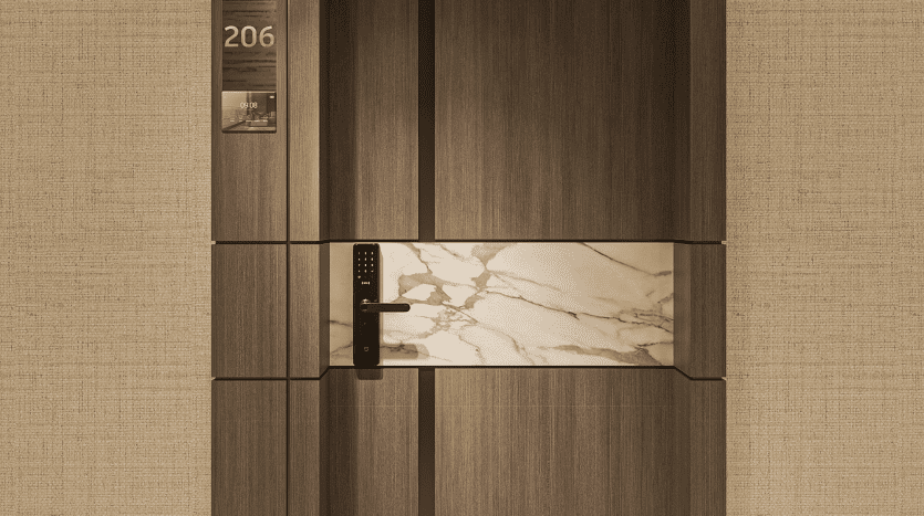 Une élégante porte en bois avec une serrure numérique et des accents de marbre, marquée du numéro de chambre 206, se dresse bien en vue dans les résidences Primero Al Furjan. Adjacent à la porte se trouve un panneau transparent montrant un petit calendrier et divers objets. Le mur entourant la porte présente un design beige texturé.