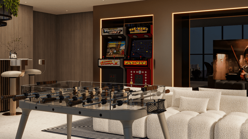Dans Primero Residences Al Furjan, une chambre moderne dispose d'une table de baby-foot en verre, d'un canapé blanc confortable, d'un tapis rayé et d'un espace bar avec des tabourets hauts. Deux machines de jeux d'arcade et un écran de télévision affichant une scène de film en arrière-plan ajoutent une ambiance de divertissement à l'espace.