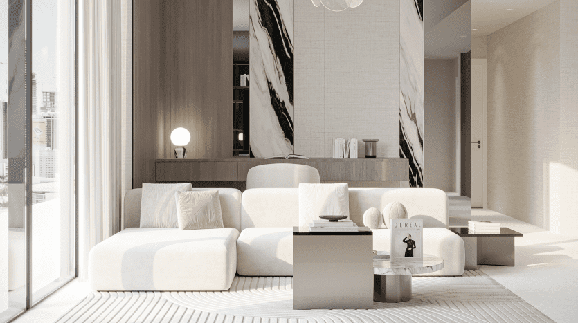 Un salon moderne et minimaliste du Primero Residences Al Furjan comprend un canapé sectionnel blanc, un tapis de couleur claire et des tables basses élégantes. Un mur d'accent en marbre avec un motif géométrique ajoute de la sophistication, tandis qu'une baie vitrée inonde l'espace de lumière naturelle.