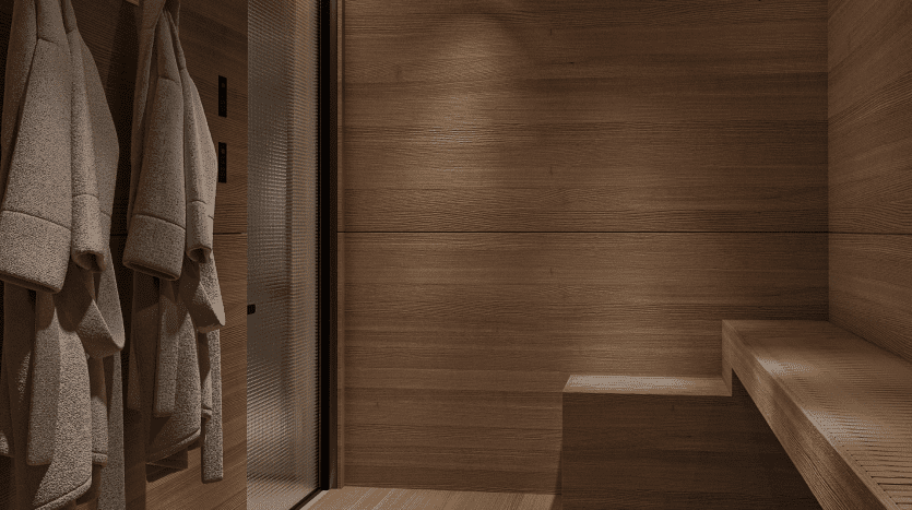 Un sauna moderne en bois du Primero Residences Al Furjan présente des murs en bois lisses marron clair et un banc assorti sur un côté. Trois peignoirs gris sont suspendus à des crochets sur le côté gauche de la pièce. L'éclairage crée une atmosphère chaleureuse et tranquille.