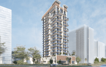 Un immeuble résidentiel moderne de grande hauteur, Primero Residences Al Furjan, doté de nombreux balcons, se dresse sur un ciel partiellement nuageux. La zone environnante comprend des bâtiments de grande hauteur supplémentaires, des arbres et un véhicule qui passe sur la route devant la structure.