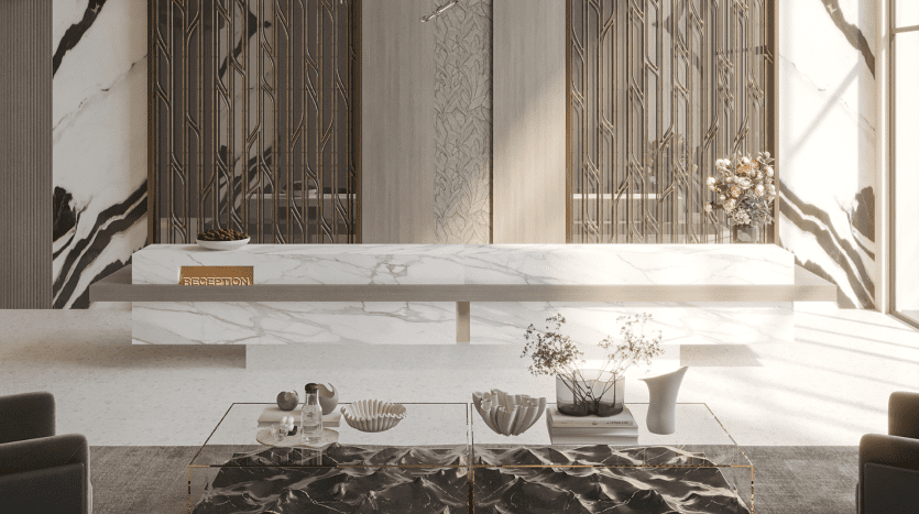La réception du Primero Residences Al Furjan comprend un bureau de réception en marbre blanc avec une signalisation dorée. Derrière le bureau se trouvent des panneaux décoratifs à motif géométrique. L'espace est agrémenté d'un bol de pommes vertes et d'un arrangement floral, tandis qu'une table basse en verre se trouve au premier plan.