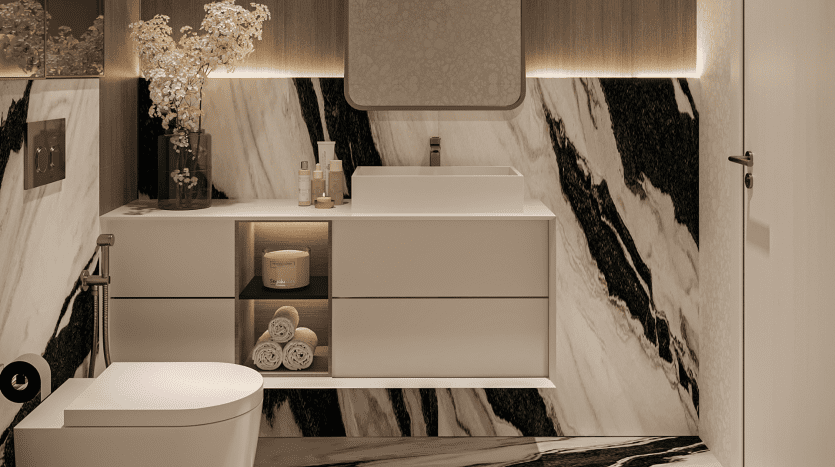 Une salle de bains moderne du Primero Residences Al Furjan présente des murs et des sols en marbre noir et blanc, avec un lavabo rectangulaire blanc sur une vanité flottante. Les étagères ouvertes sous l'évier contiennent des serviettes roulées et des articles de toilette. Un miroir rétroéclairé est suspendu au-dessus, tandis que des toilettes sont visibles sur la gauche.