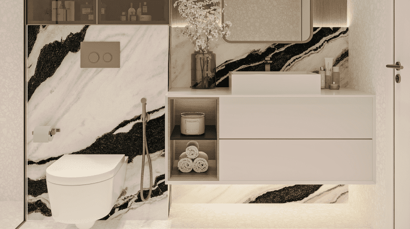 La salle de bains moderne du Primero Residences Al Furjan présente des murs et un sol en marbre crème et noir. Une vanité flottante blanche avec un lavabo au-dessus du comptoir, des articles de toilette et des objets décoratifs se trouve à droite. À gauche se trouvent des toilettes suspendues blanches avec un bec de bidet et un porte-papier toilette.
