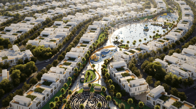 Vue aérienne d'un quartier résidentiel moderne du sud de Dubaï avec de nombreux bâtiments blancs, une verdure luxuriante et un lac central entouré de palmiers et de sentiers pédestres. Aire de jeux centrale colorée et jardins paysagers visibles.