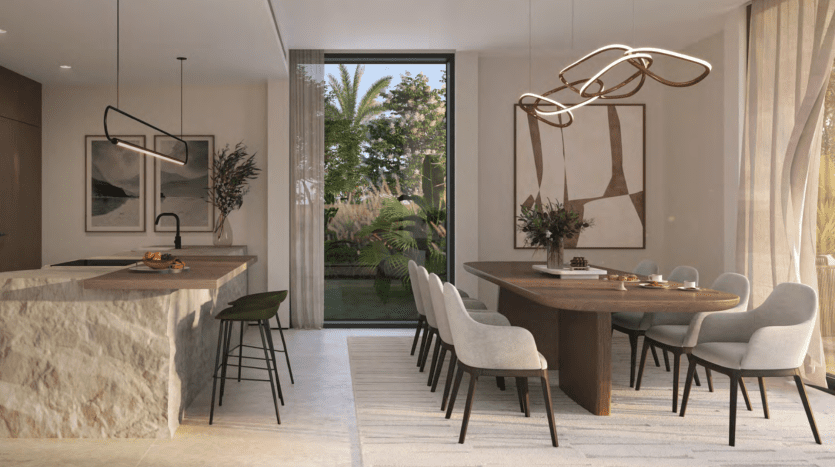 Une salle à manger moderne dans une villa avec une table ovale en bois entourée de chaises grises, adjacente à une cuisine avec un îlot en marbre et des tabourets de bar. De grandes fenêtres affichent un jardin luxuriant. Luminaires dorés élégants