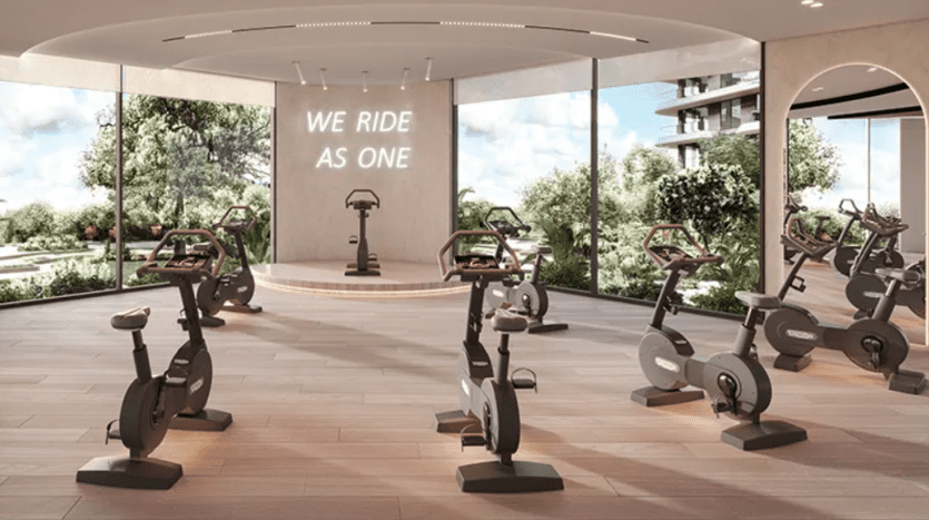 Un studio de cyclisme en salle moderne avec plusieurs vélos stationnaires disposés face à une plate-forme surélevée sur laquelle repose un seul vélo. Un panneau sur le mur du fond indique « WE RIDE AS ONE ». De grandes fenêtres offrent une vue sur la verdure luxuriante à l'extérieur et la pièce est dotée de parquet.