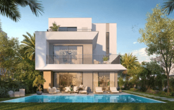 Une maison blanche moderne de deux étages dotée de grandes fenêtres et de balcons est nichée le long d'une ruelle calme, entourée d'une verdure luxuriante. Dans la cour, il y a une piscine rectangulaire avec des chaises longues et des plantes autour du patio. Le ciel est clair et bleu.