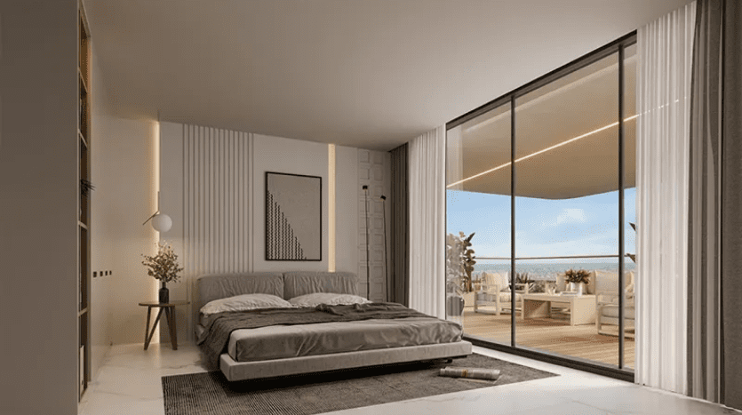Une chambre moderne avec une grande porte vitrée coulissante du sol au plafond menant à un balcon. La chambre comprend un lit double avec une literie grise, une table de chevet avec une lampe, des rideaux du sol au plafond, une œuvre d'art murale et une vue sur le mobilier d'extérieur sur le balcon.