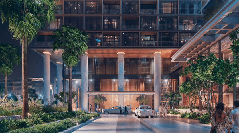 Une entrée d'hôtel moderne et éclairée la nuit avec de hautes fenêtres et des piliers en verre respire la verve. Trois voitures sont garées devant l’entrée et des gens marchent près de la porte. La zone est entourée de verdure, comprenant des palmiers et des buissons.