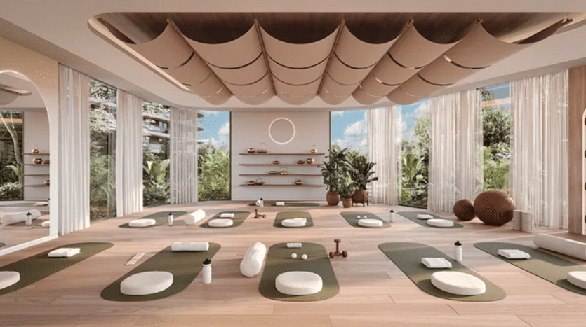 Un studio de yoga spacieux et bien éclairé avec de grandes fenêtres et une douce lumière naturelle comprend des tapis, des traversins et des accessoires soigneusement disposés sur un plancher en bois. Des rideaux légers et fluides encadrent les fenêtres, tandis que les plantes vertes vertes à l'extérieur ajoutent une atmosphère sereine.
